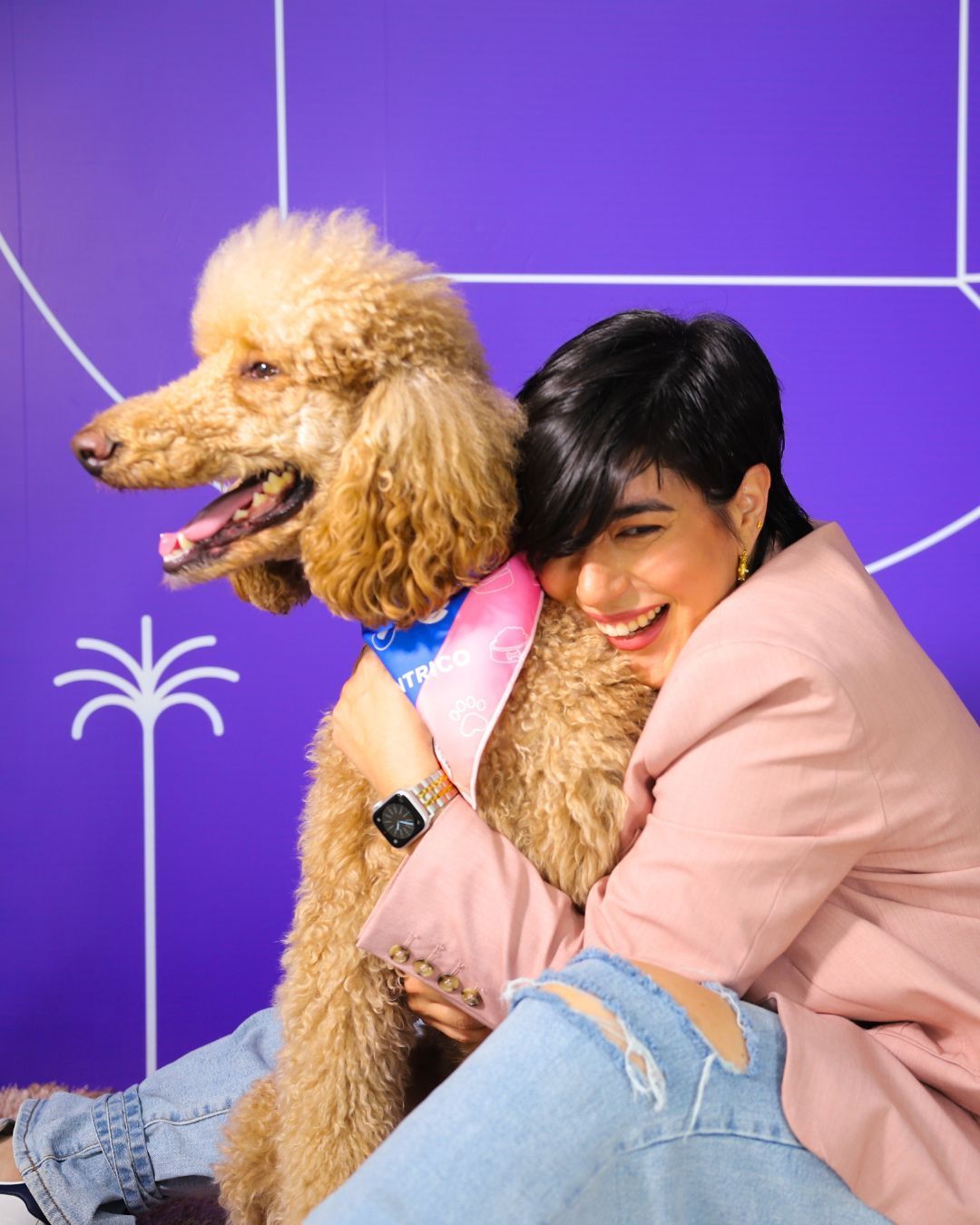 Woman with short dark hair hugs a tan poodle, both smiling in front of a purple background.