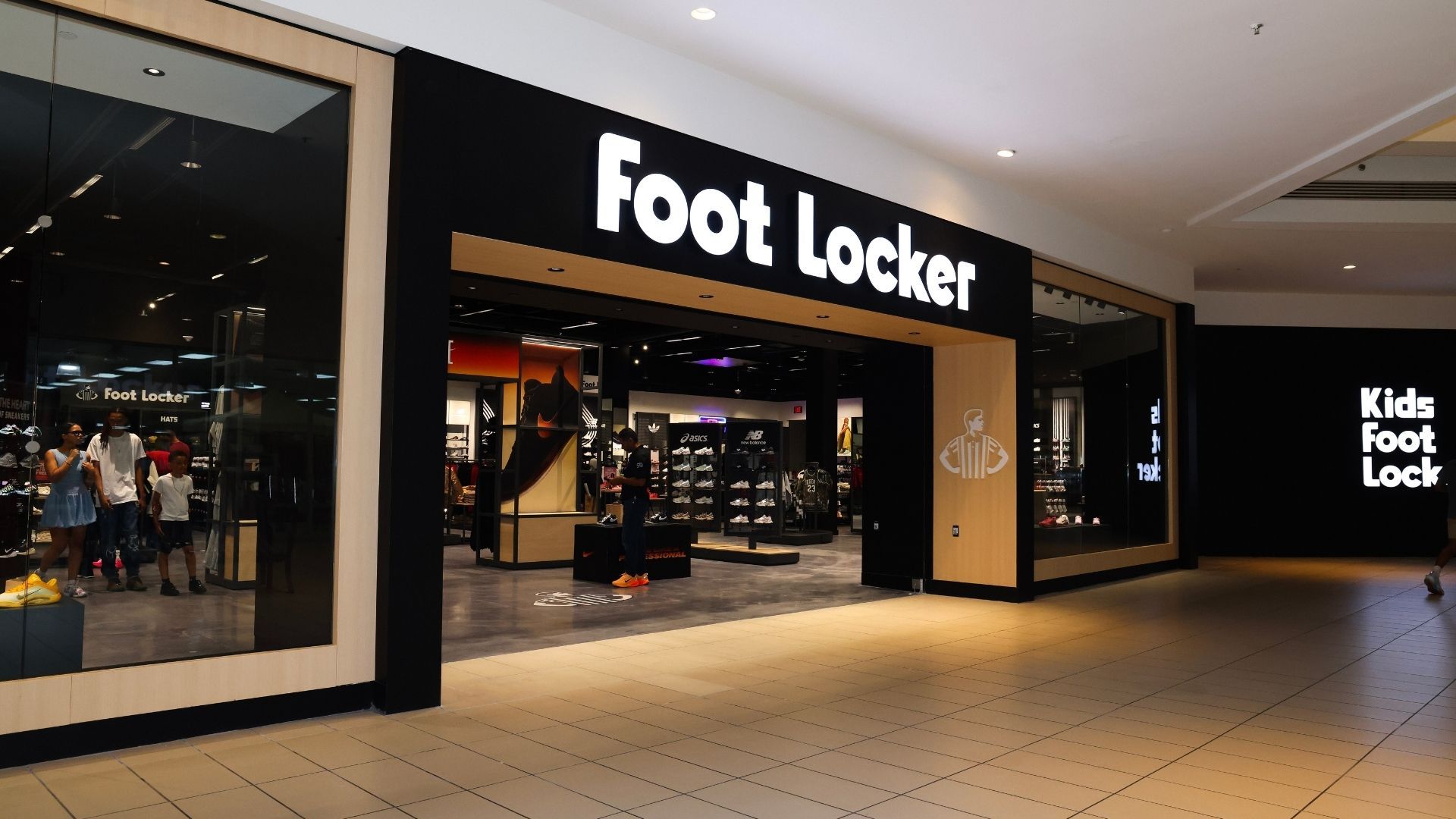 Foot Locker store entrance in a mall, black and white signage, beige trim, glass windows, with some people inside.