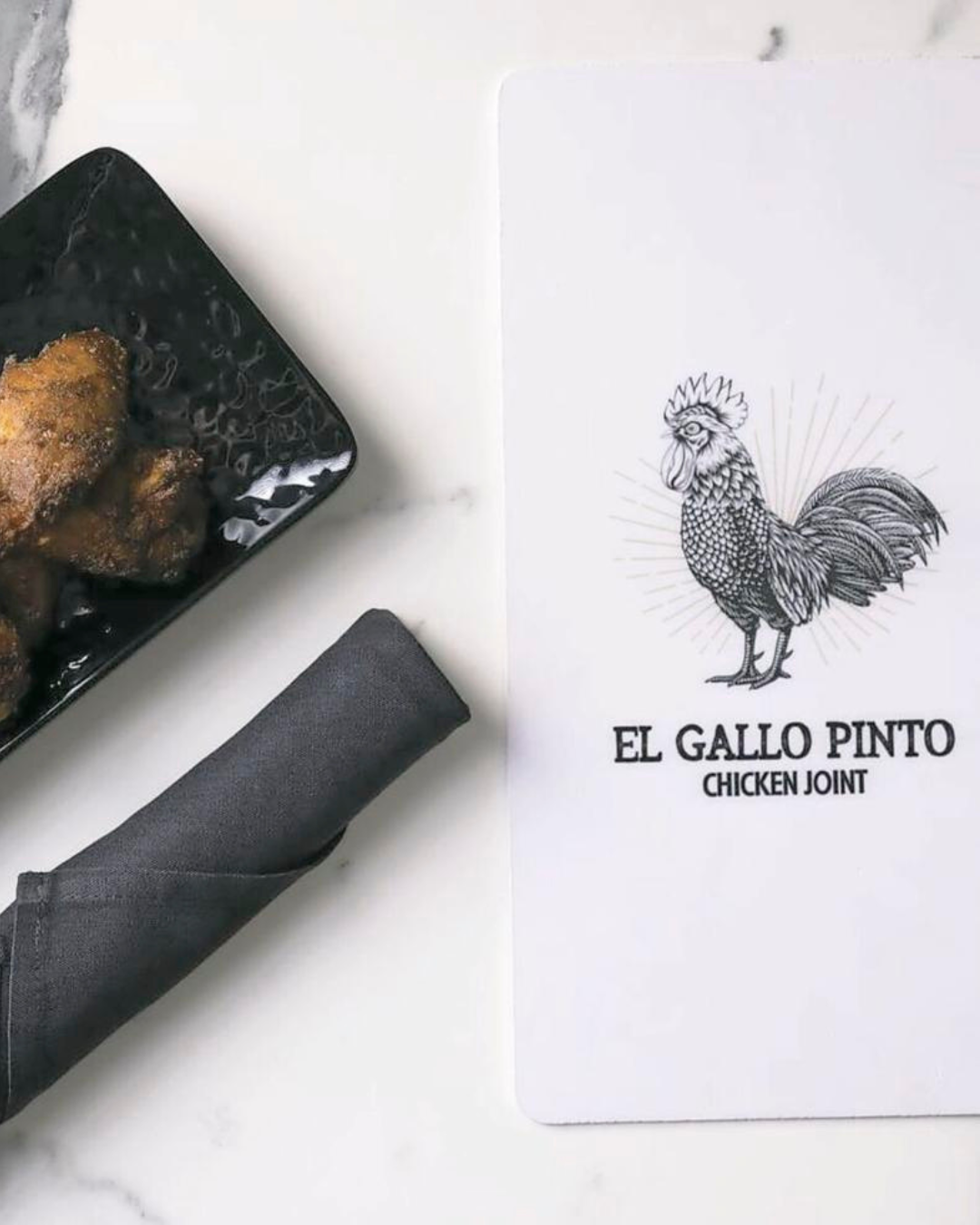 Black plate with fried food, rolled napkin, and menu with rooster illustration and