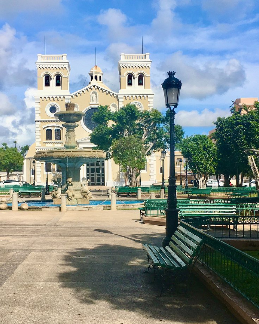 Plaza with fountain, benches, and church with two towers under a partly cloudy sky.