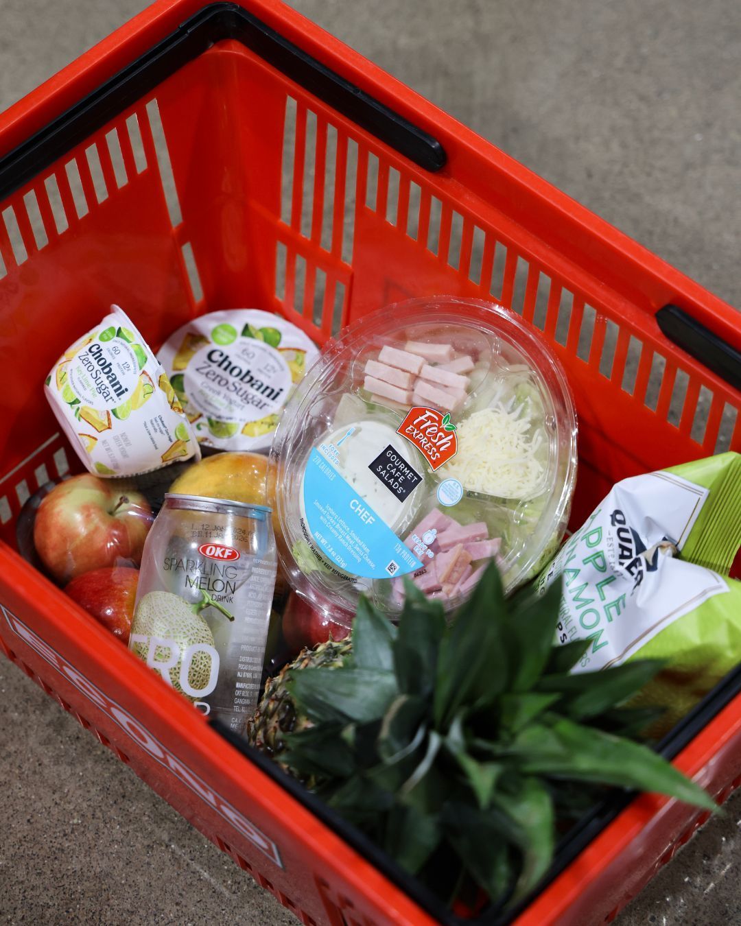 Red shopping basket filled with cheese, fruit, and snacks.