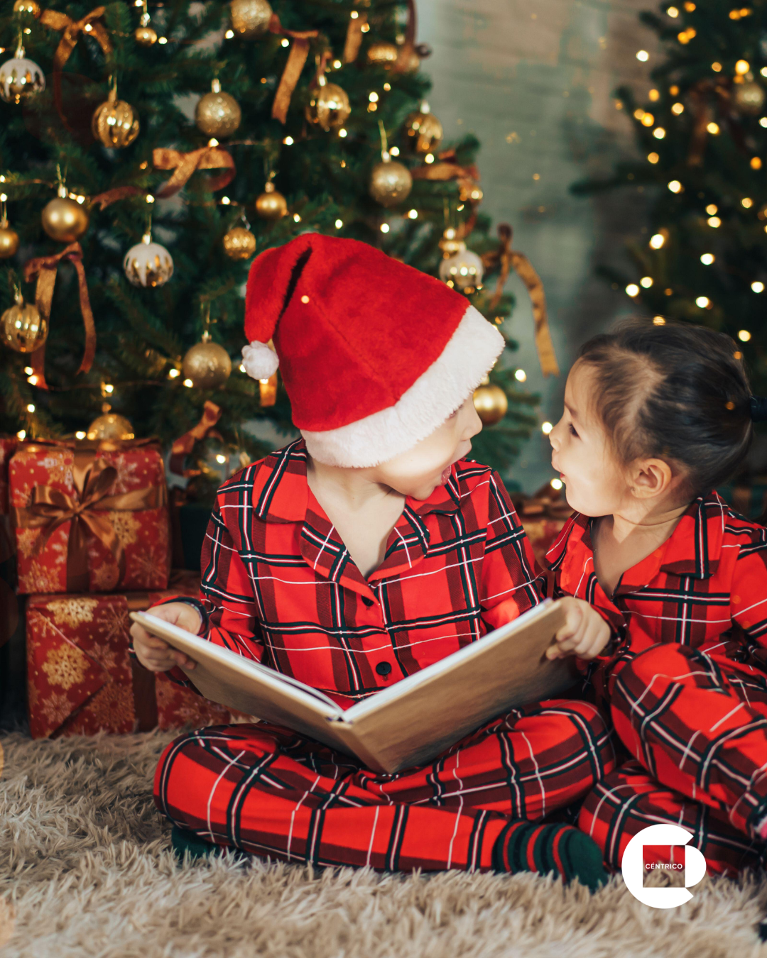 Two children in matching plaid pajamas and a Santa hat read a book near a Christmas tree.