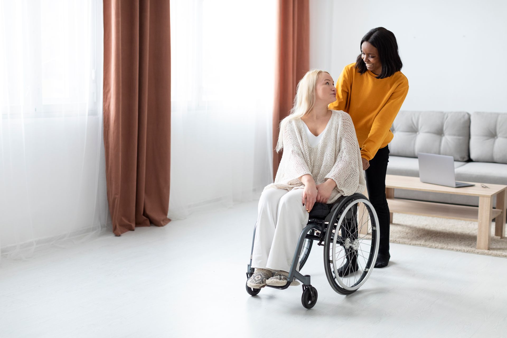 Friendly NDIS disability support worker pushing smiling woman in wheelchair in bright, accessible living room
