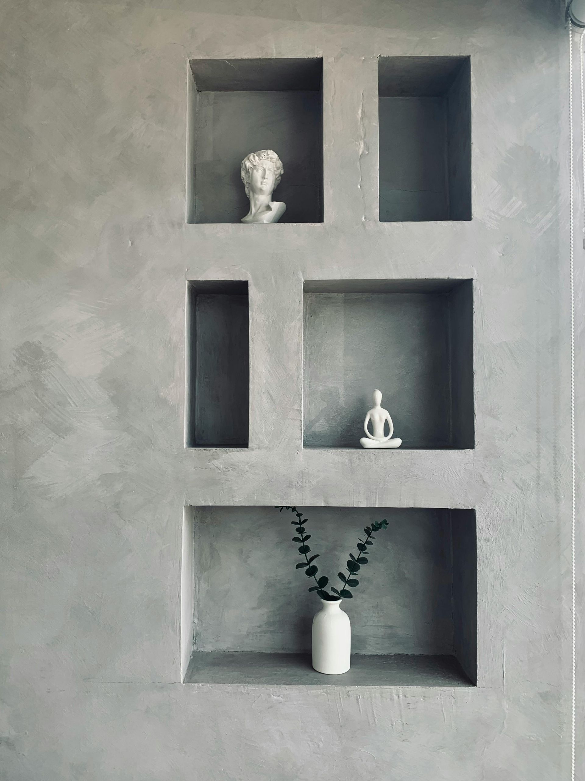 Gray wall with built-in square and rectangular niches, displaying a bust, a seated figure, and a vase with greenery.