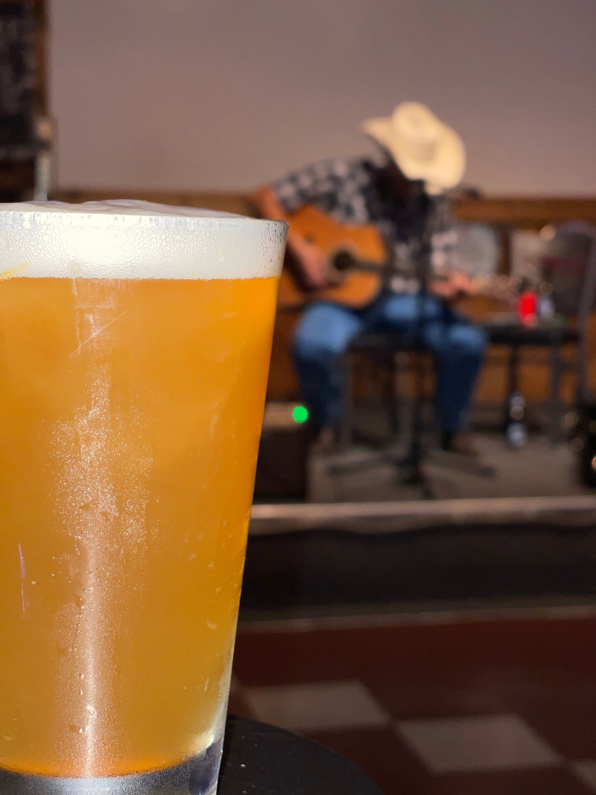 A close up of a glass of beer with a man playing a guitar in the background.