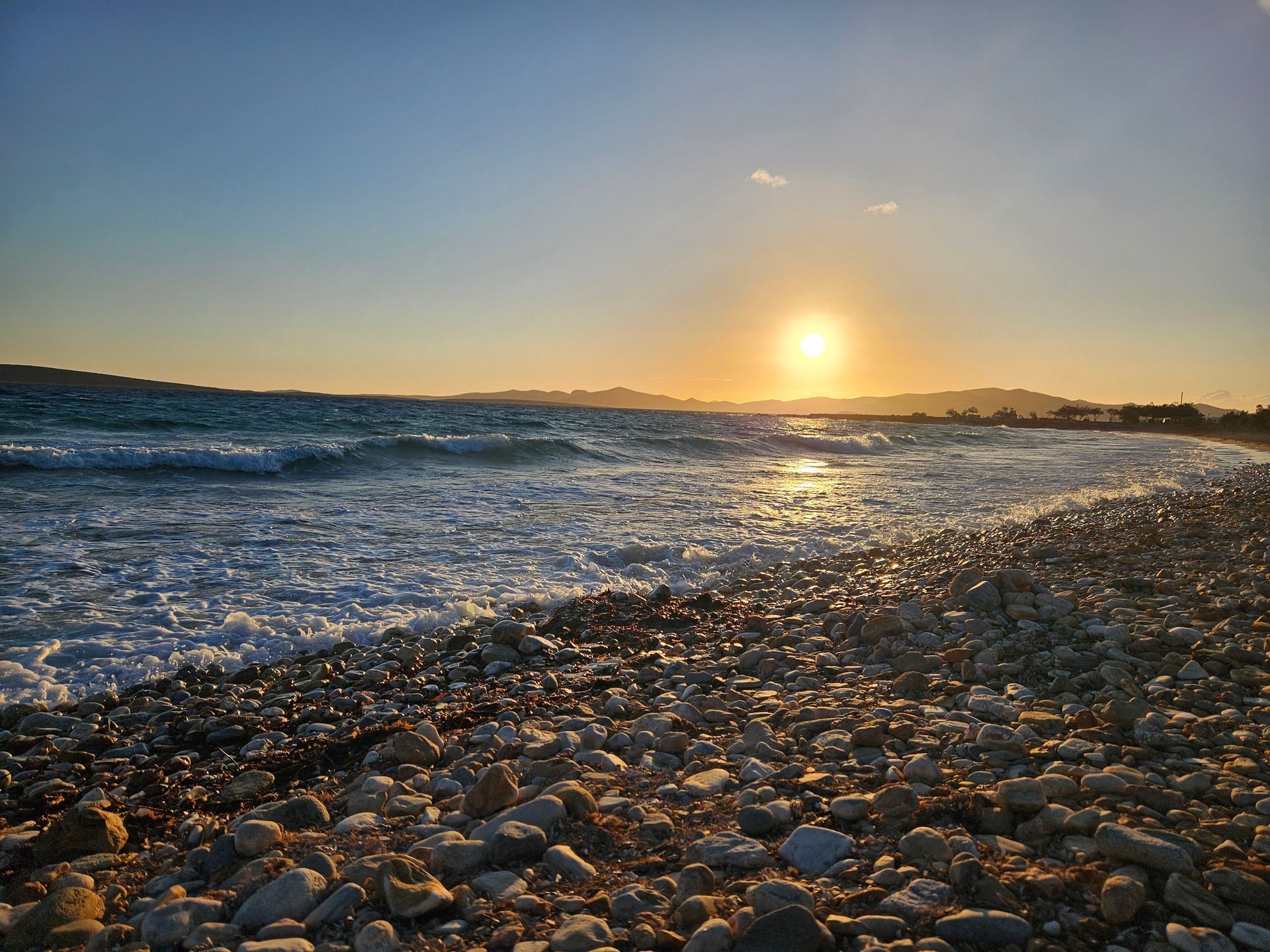 Sunset over ocean waves crashing on a pebbled beach. Warm sunlight reflects on water and stones.