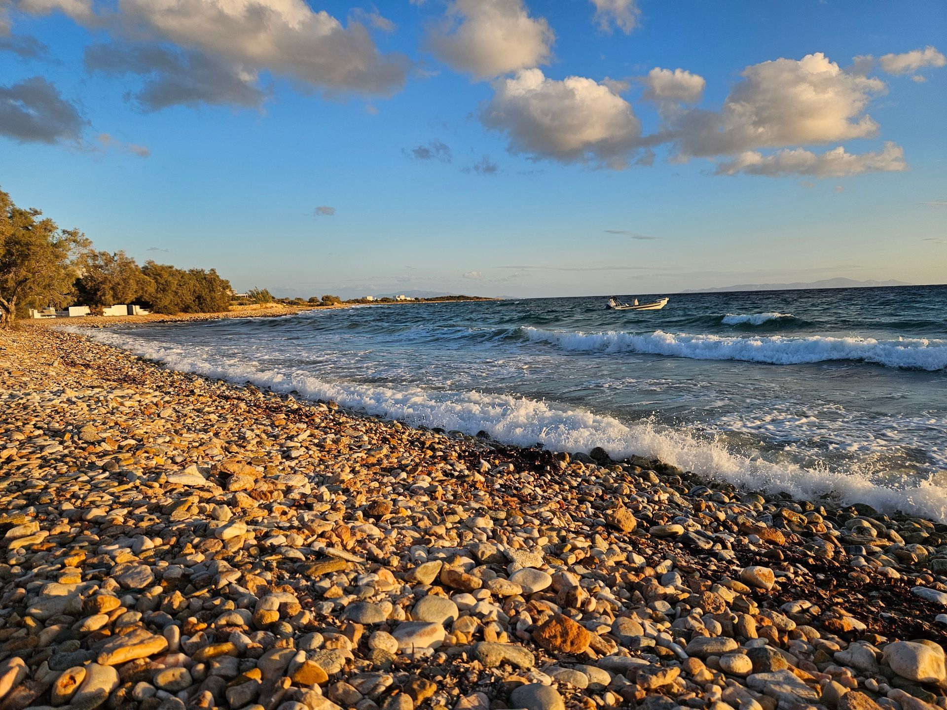 Pebble beach with waves crashing. Blue sky, white clouds, small boats in the distance, and golden sunlight.