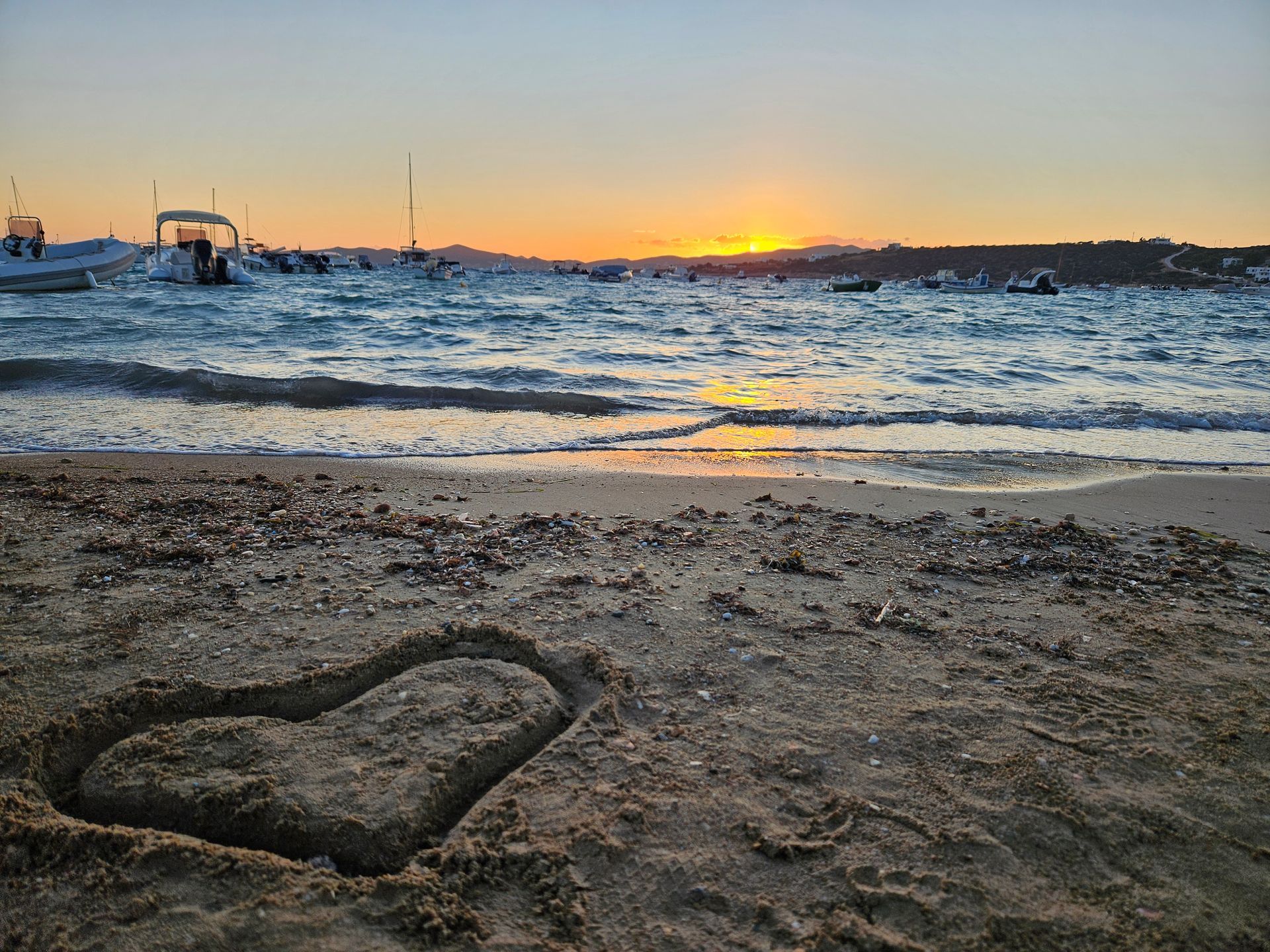 Heart shape drawn in sand on a beach at sunset; boats on the water.