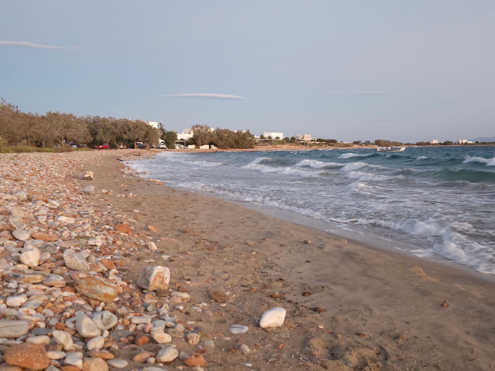 Beach with sand and pebbles, waves, blue water, and buildings along the shore.