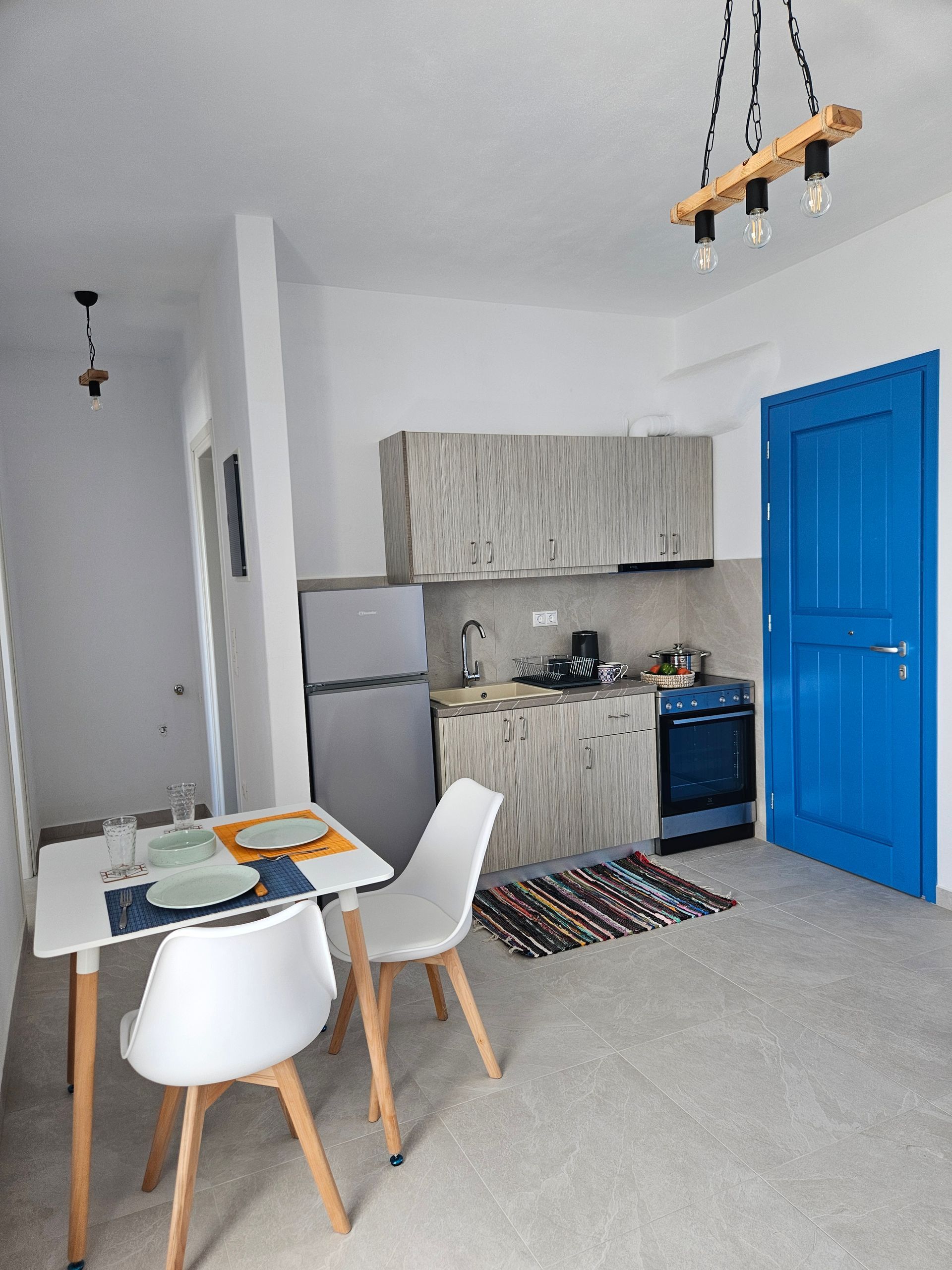 Small studio kitchen and dining area with white walls, blue door, and simple furniture.
