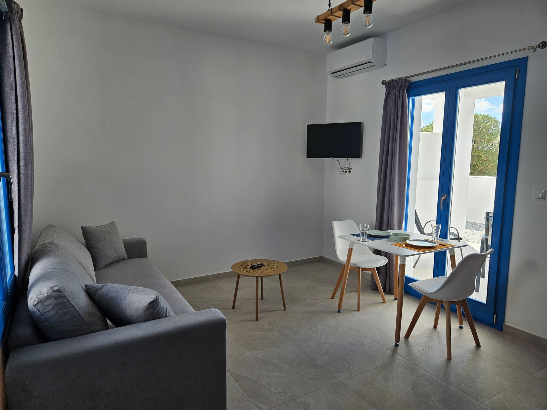 A modern, bright living space with a grey sofa, small table, dining table and chairs, and a blue-framed door.