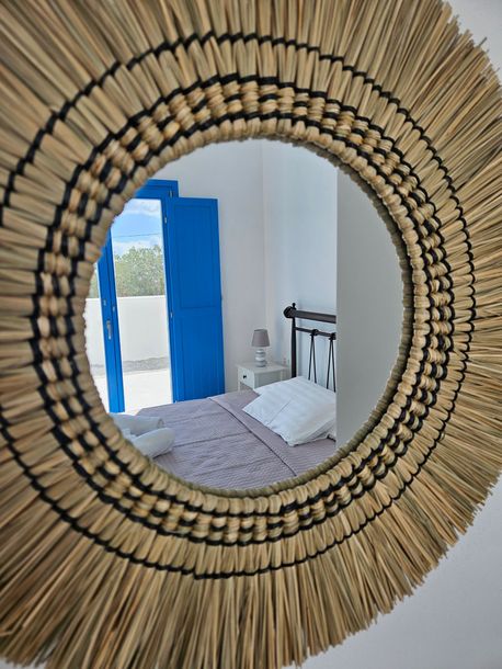 Round straw mirror reflecting a bedroom with a blue door, bed, and white walls.