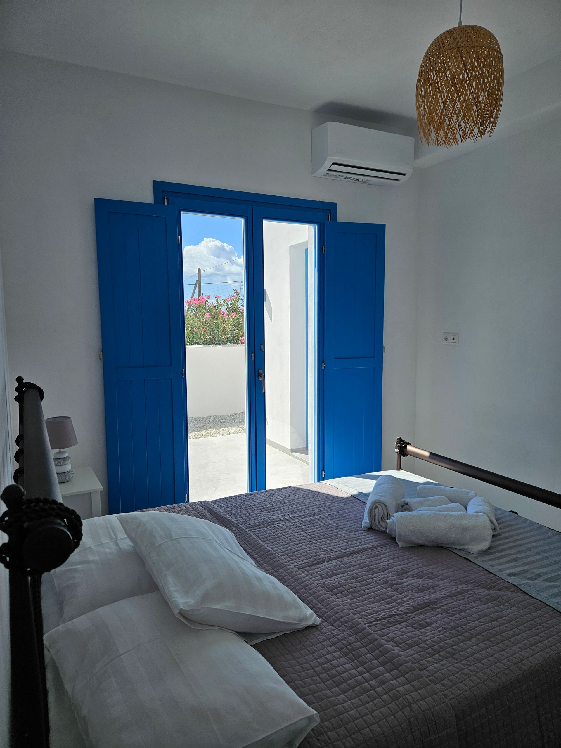 Bedroom with blue doors leading to a balcony. White walls, grey bedding, and a wicker lamp.