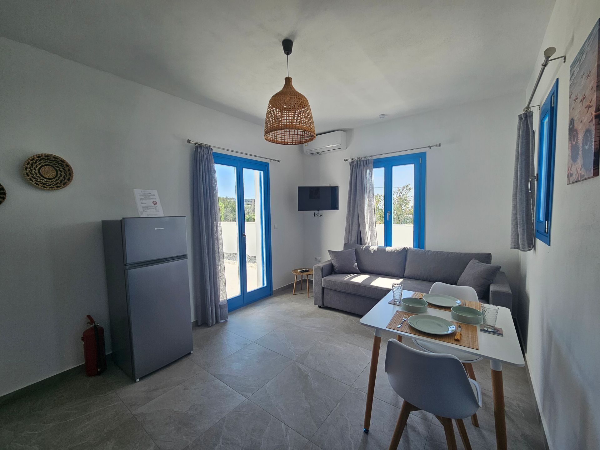 Living room with blue window frames, gray sofa, fridge, and rattan pendant lamp.