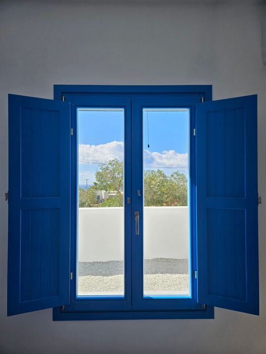 Blue shutters open on a window, revealing blue sky and a white wall, possibly in Greece.