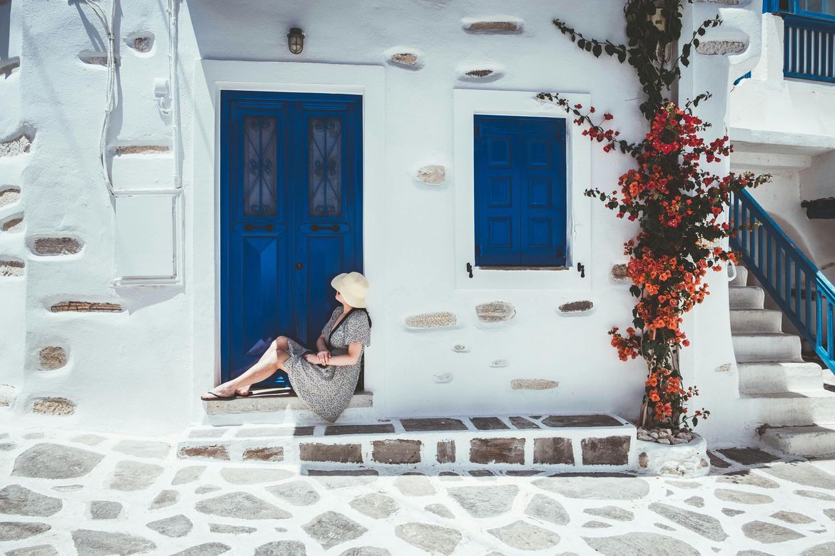 Woman sits on steps of a white building with blue door and window, red flowering vine, and stone pavement.