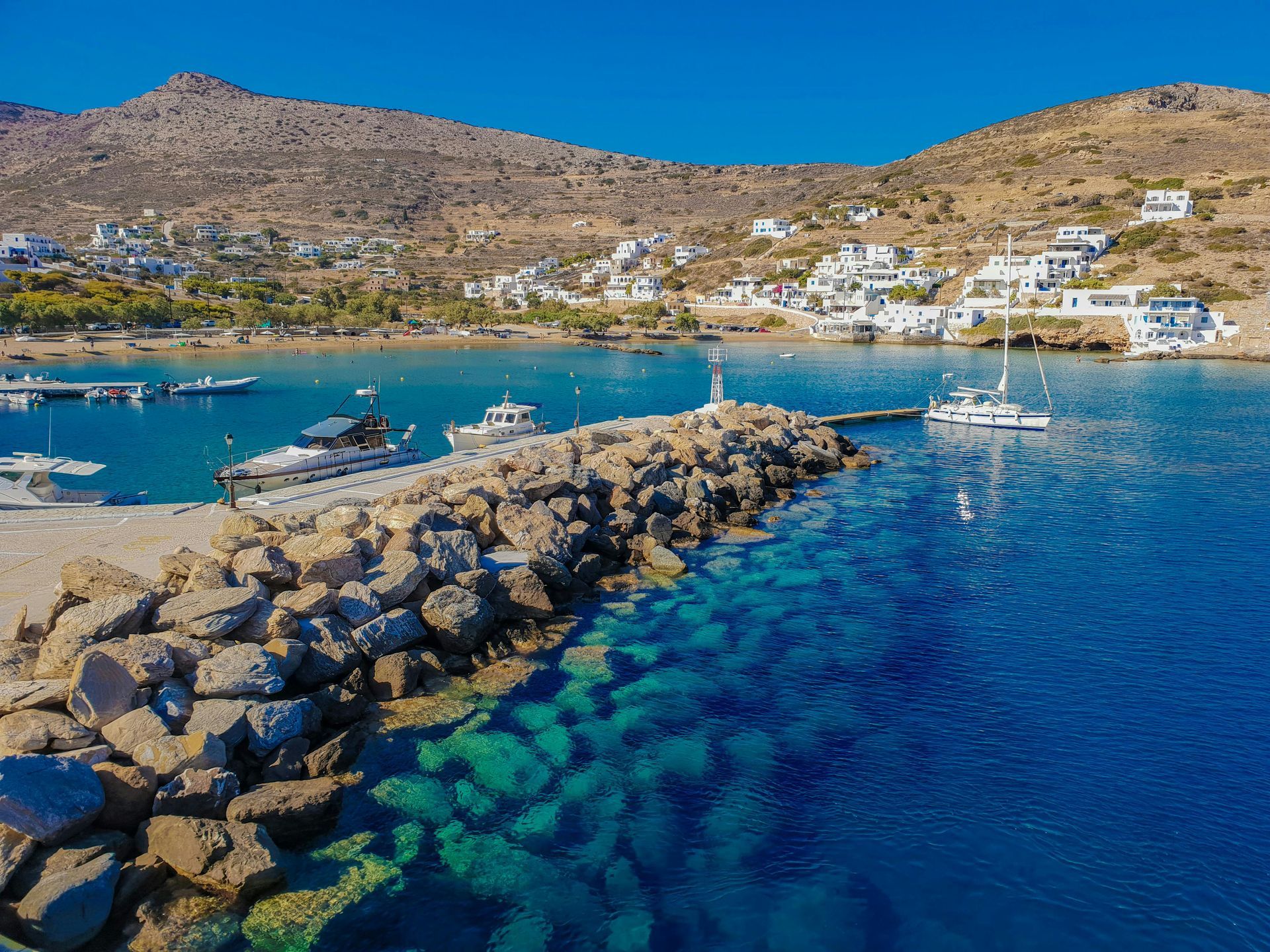 Rocky pier with boats on turquoise water, white buildings on a hillside under a blue sky.