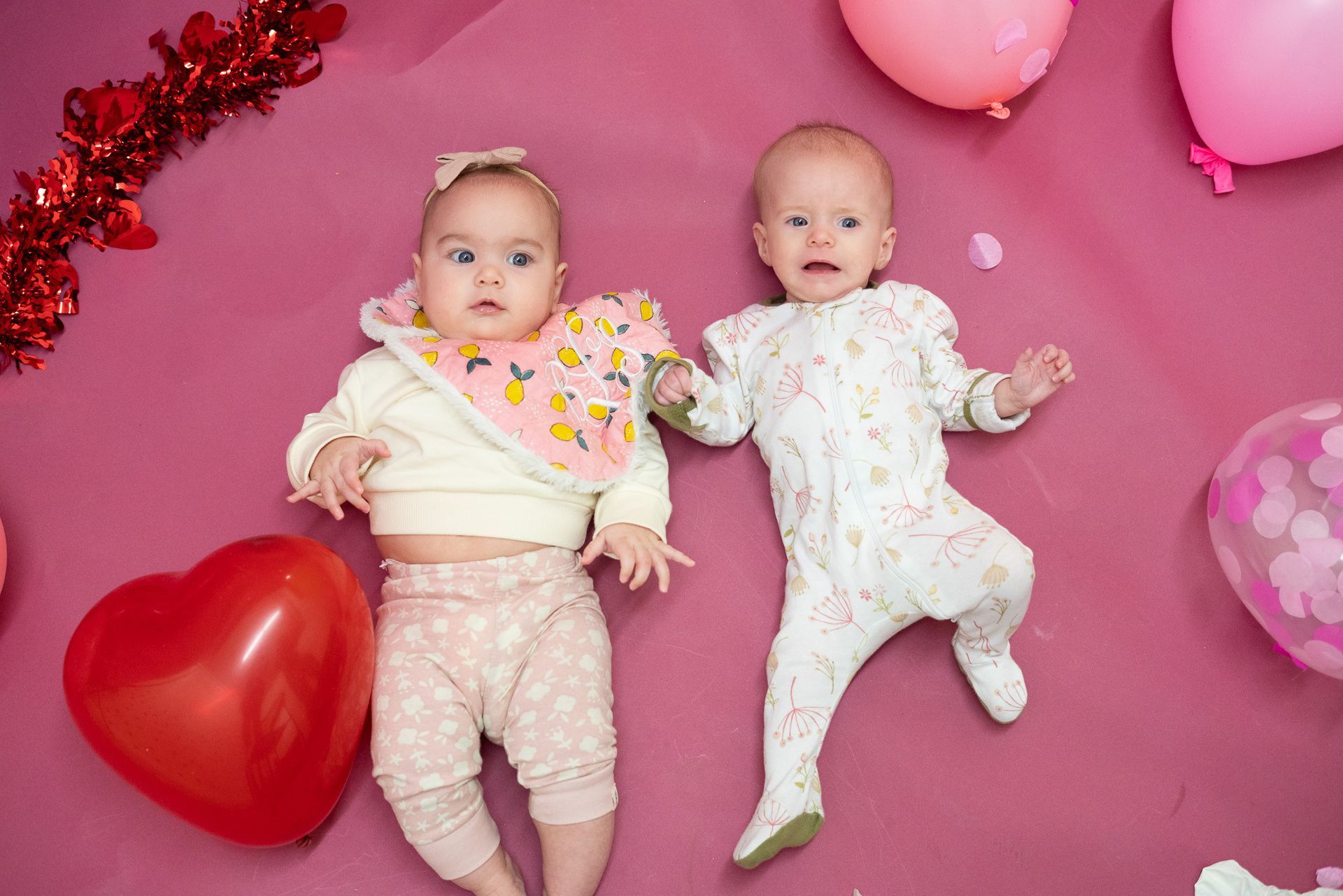 Two babies are laying next to each other on a pink surface surrounded by balloons.