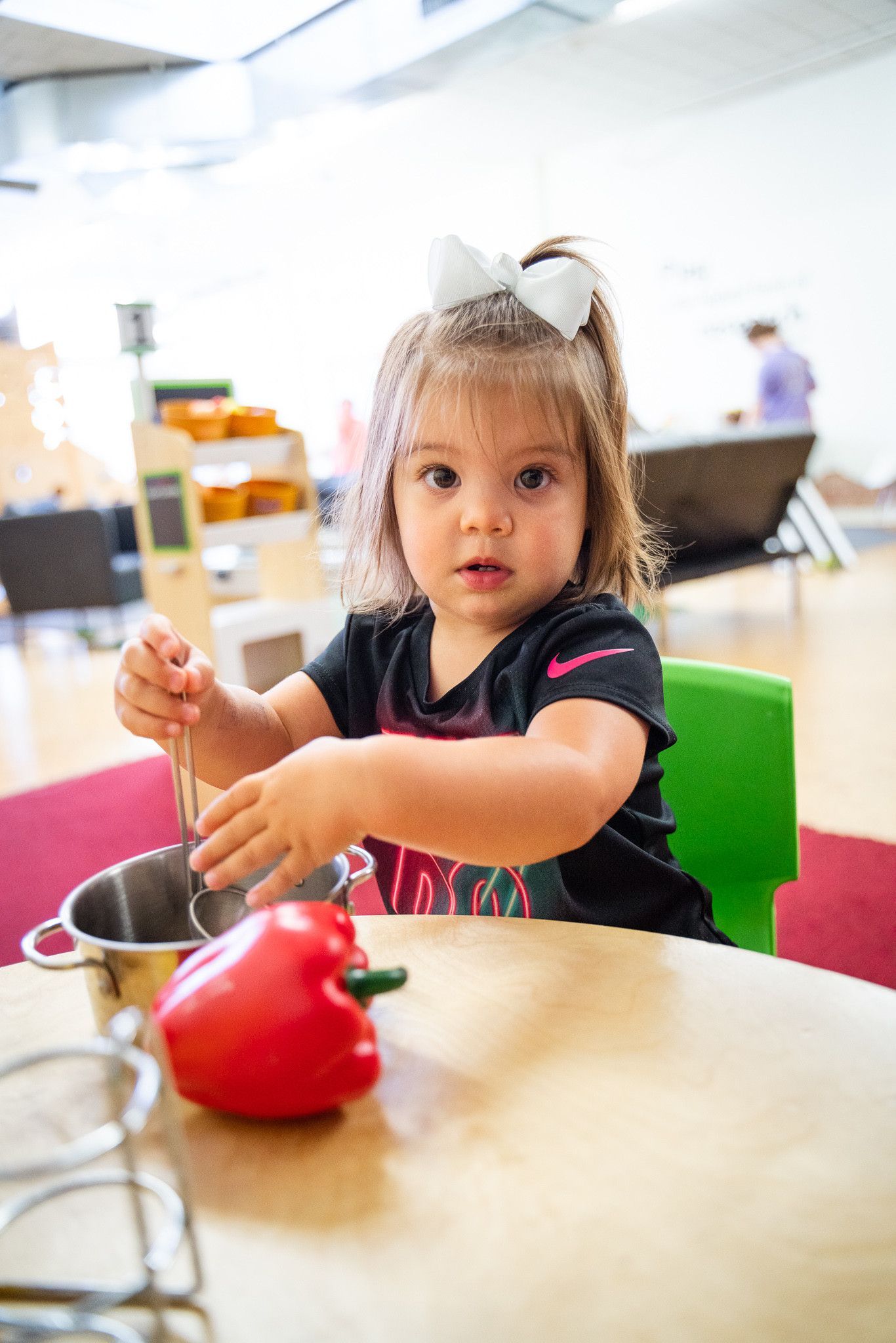 A little girl is sitting at a table playing with a red pepper.