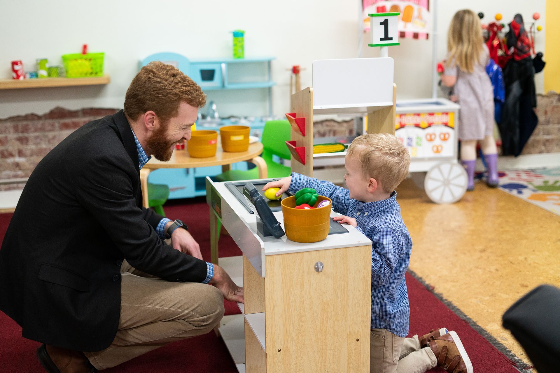 A man is kneeling down next to a little boy playing with a toy kitchen.