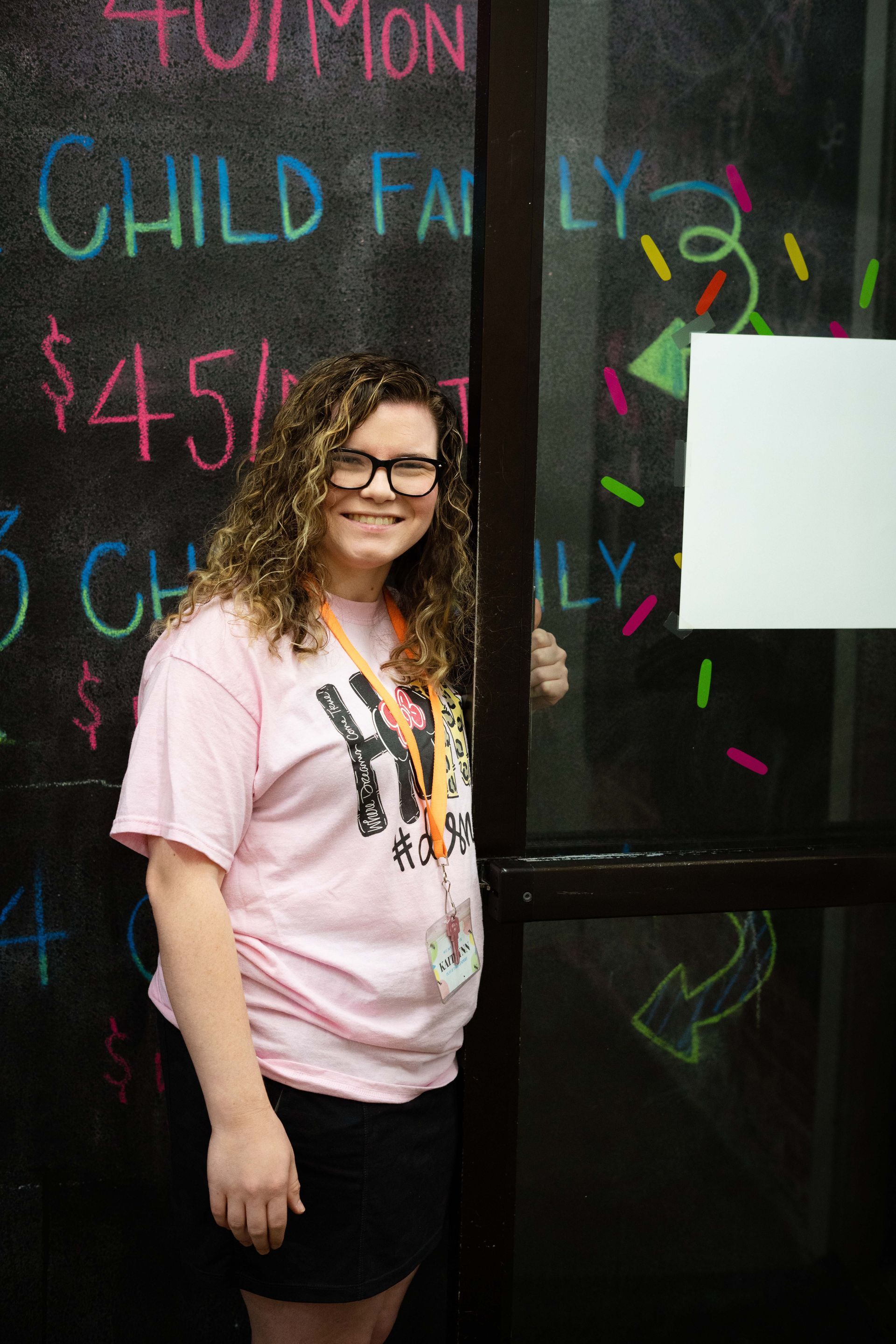 A woman in a pink shirt is standing in front of a chalkboard.