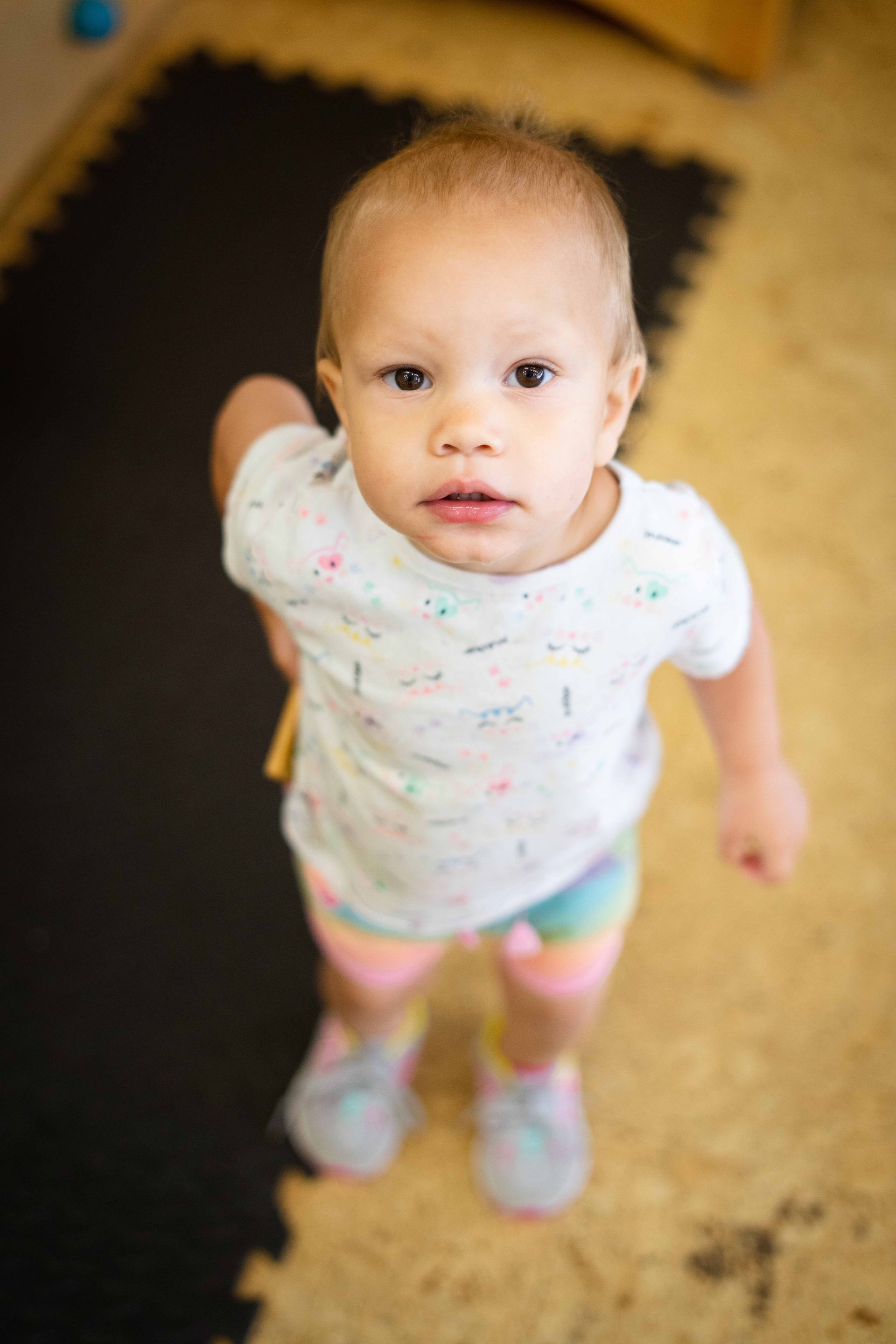 A baby girl is standing on the floor looking up at the camera.