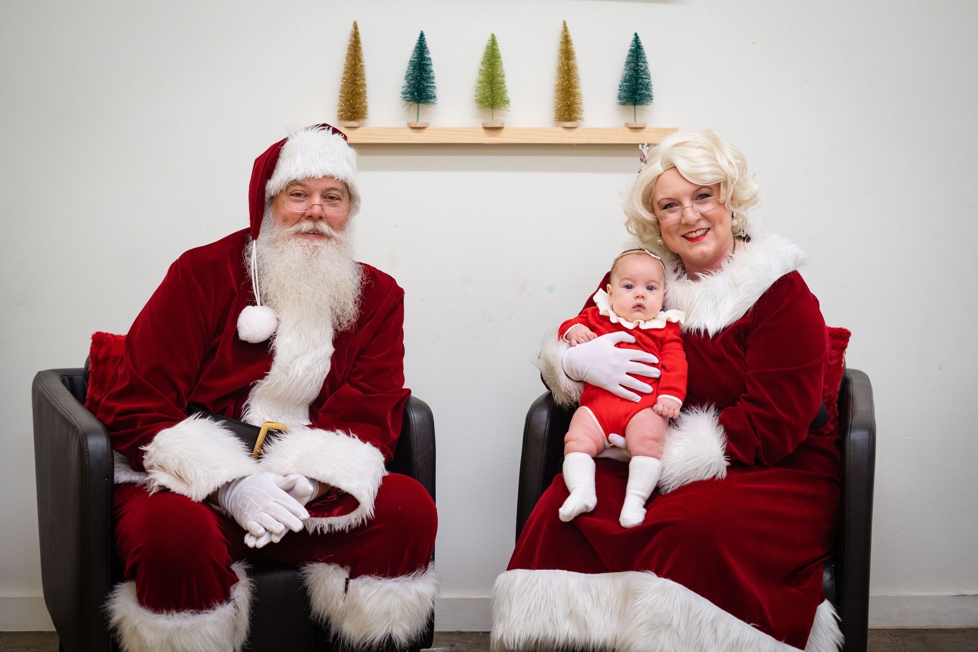 A man and a woman dressed as santa claus are sitting in chairs holding a baby.