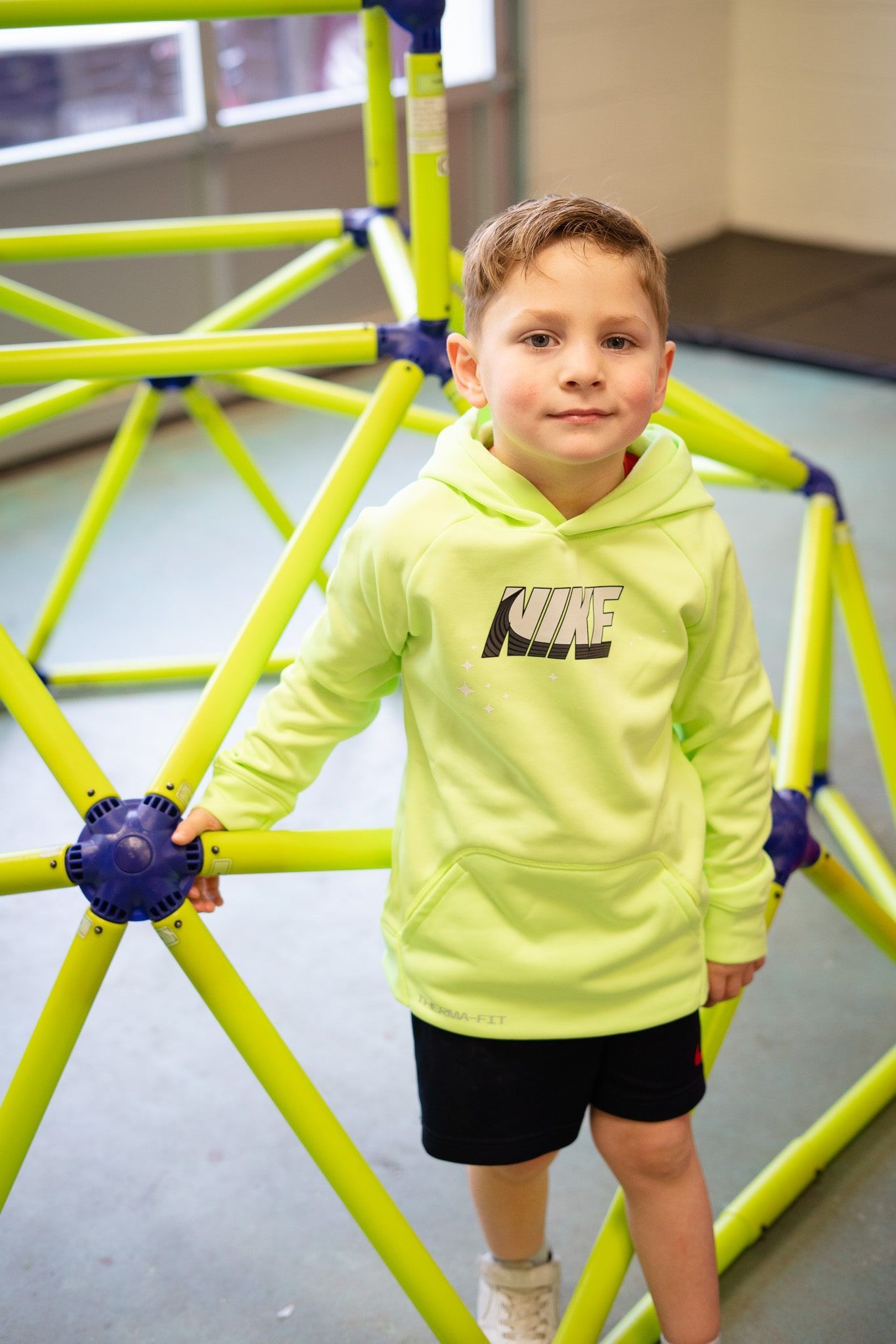 A young boy in a neon green hoodie is standing on a monkey bars.