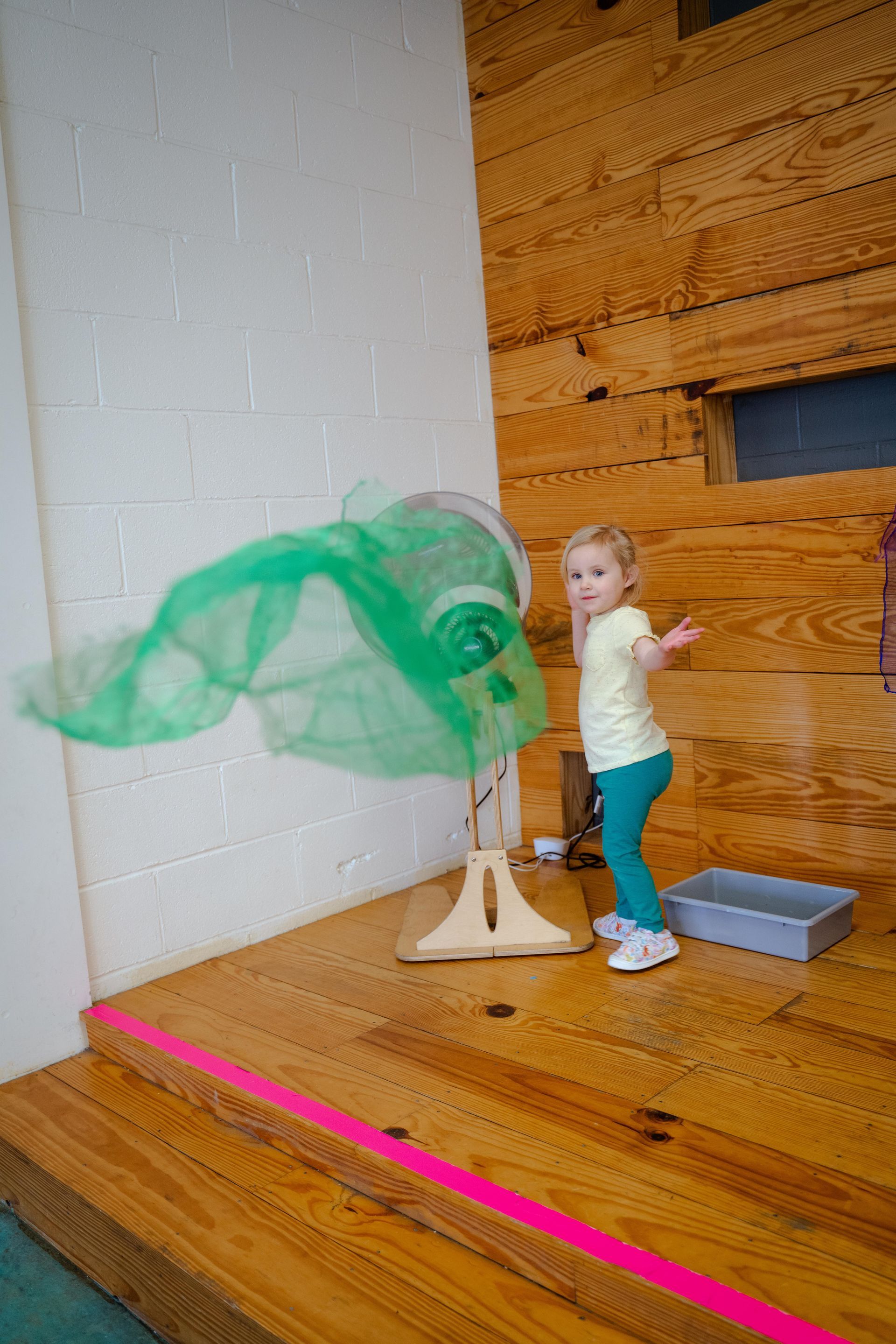 A little girl is standing next to a fan on a wooden floor.