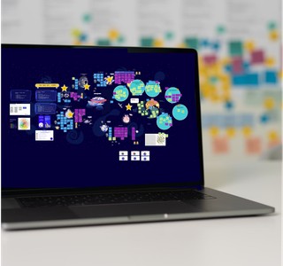 Laptop displaying a colorful graphic with text and shapes; a whiteboard with sticky notes is in the background.