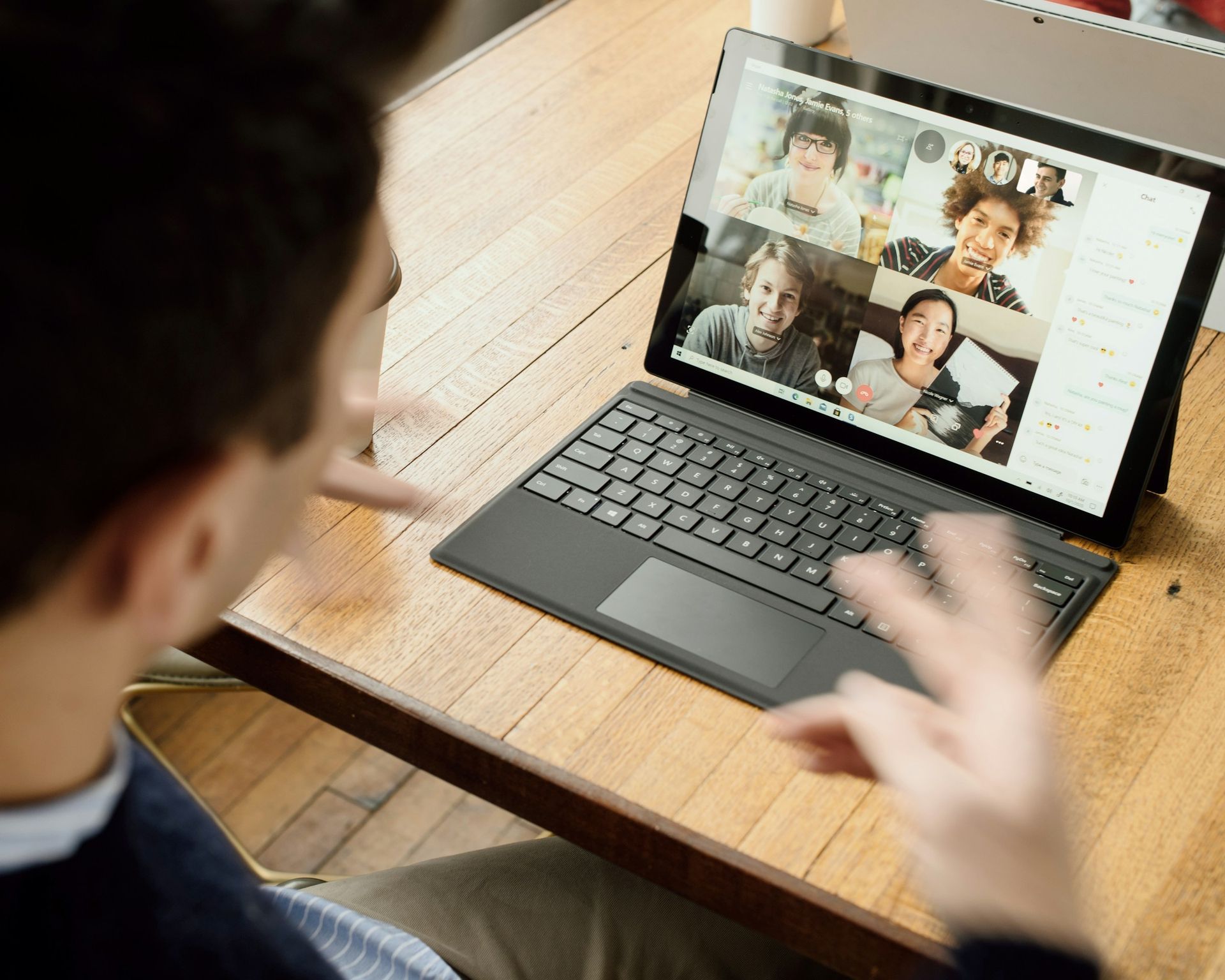 Man in video call gesturing at laptop on wooden desk with colleagues on screen.