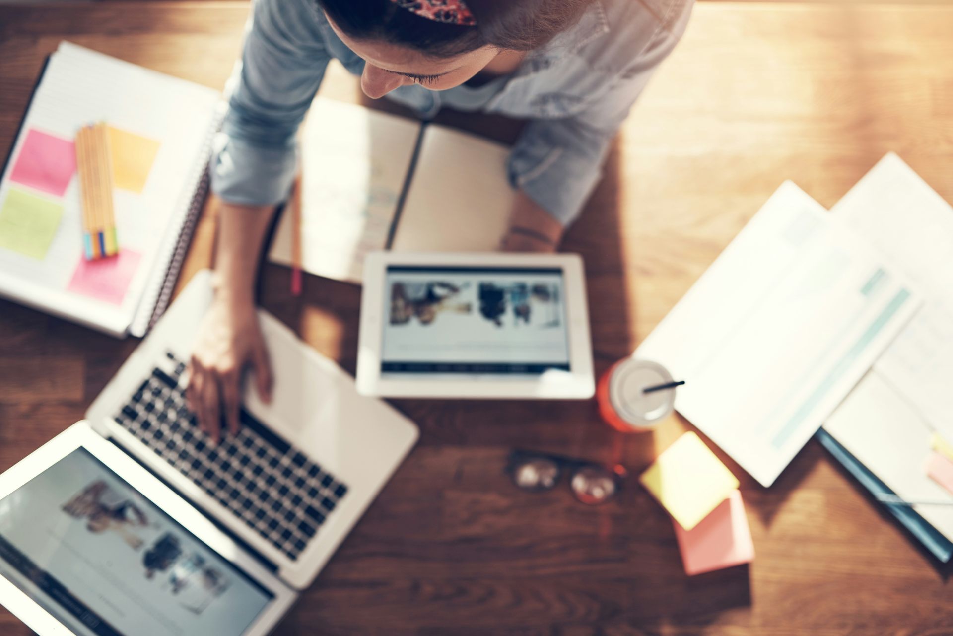 Person working at a desk with a laptop, tablet, papers, and a drink.