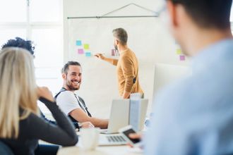 Group in a meeting, man writing on whiteboard, another smiling, others listening. Bright office.