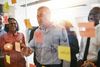 Group of diverse professionals collaborating at a glass wall covered in sticky notes; smiling and gesturing.