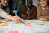 Group brainstorming, hand pointing at a pink sticky note on a white paper diagram, team smiling.