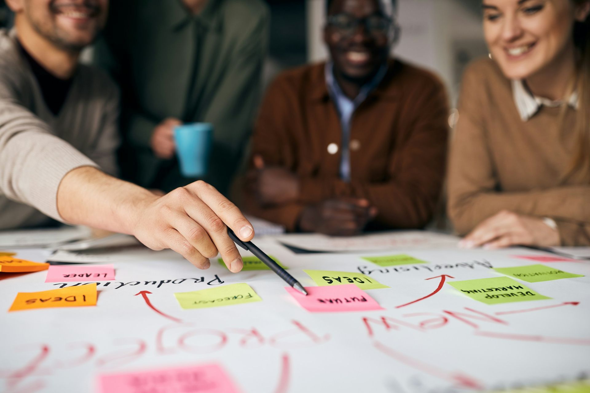 Group brainstorming, hand pointing at a pink sticky note on a white paper diagram, team smiling.