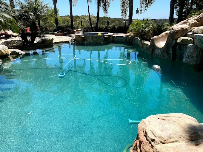Turquoise backyard pool with rock edges, palm trees, and a waterfall under a sunny blue sky