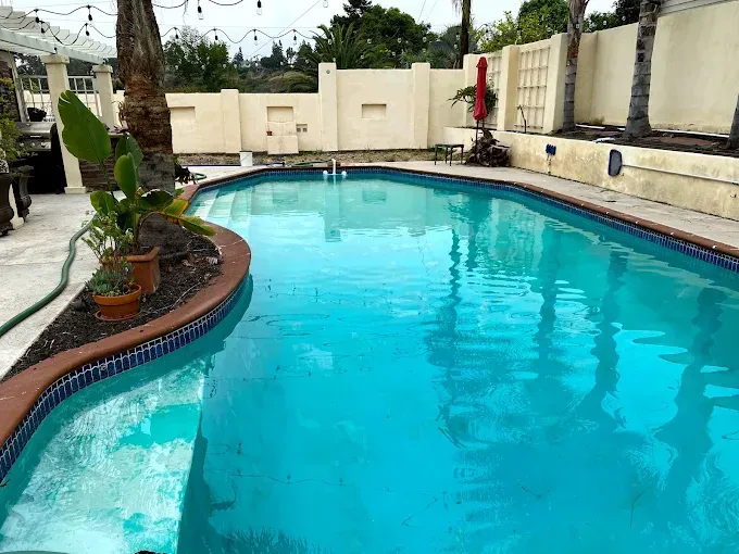 Backyard swimming pool with bright blue water, beige walls, and potted plants alongside the deck