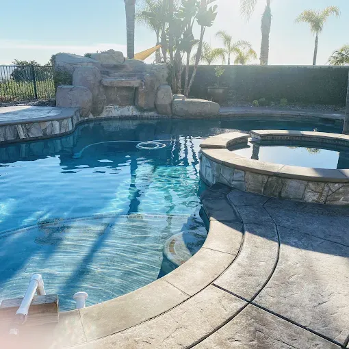 Backyard pool with rock waterfall, spa, and curved stone deck in bright sunlight