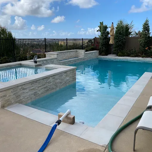 Backyard swimming pool with hot tub, blue water, stone coping, and patio hose under a partly cloudy sky