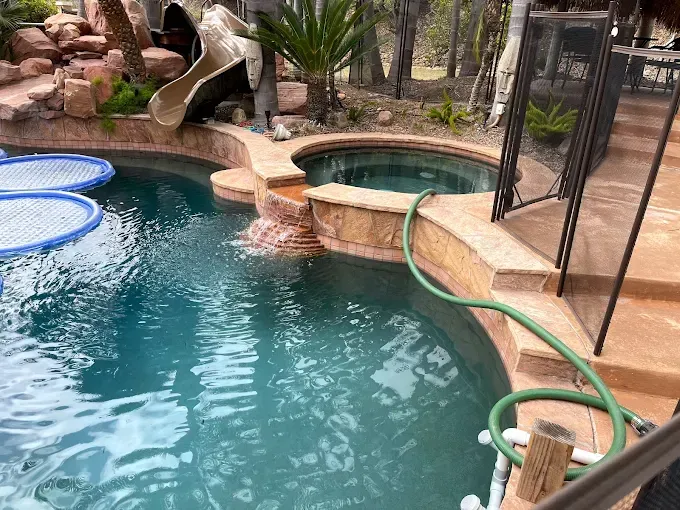 Backyard pool with blue water, stone slide, hot tub, and safety fence