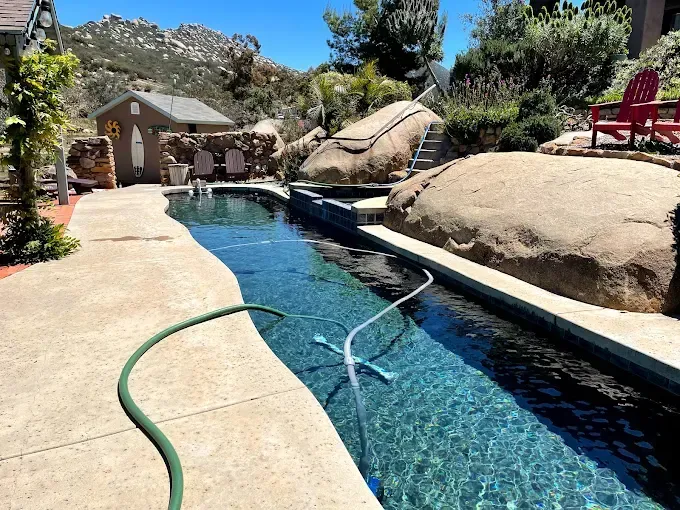 Backyard pool with clear blue water, stone deck, hose, and rocky desert landscaping