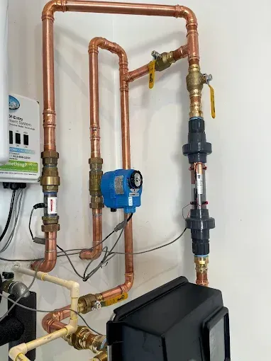Copper plumbing pipes and valves on a white wall, with a blue pump and black unit in a utility room.