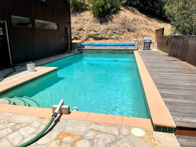 Backyard swimming pool with turquoise water, wood deck, and stone patio beside a hillside