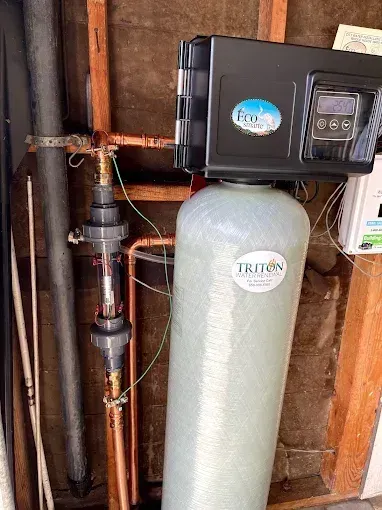Water softener system with black control unit and gray tank in a wooden utility closet.