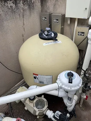 Swimming pool filter and pump equipment with beige tank and white pipes outdoors against a wall