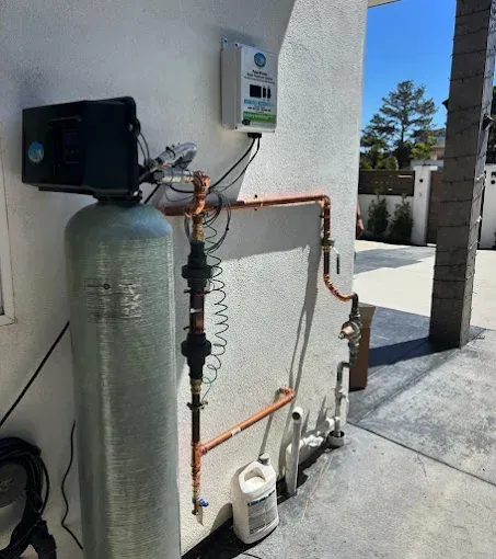 Outdoor water softener and piping setup beside a house wall with a large tank and meter.