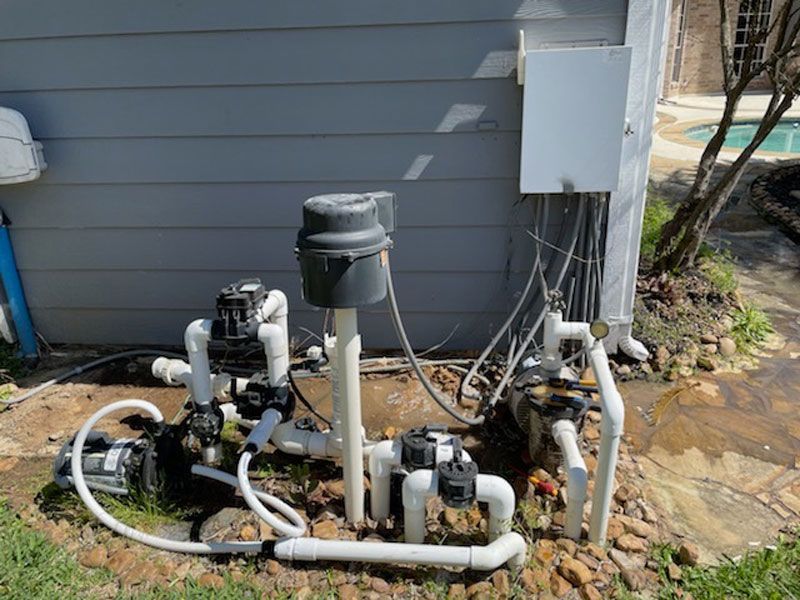 Pool equipment and plumbing beside a gray house wall, with pipes, pumps, and control box outdoors.