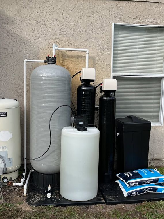 Outdoor water filtration system with tall tanks and pipes beside a house wall, near a window.