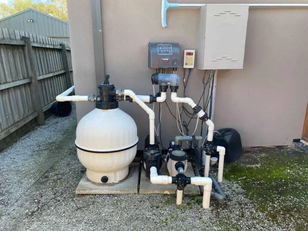 Pool filtration and pump system beside a house, with white pipes, tanks, and control units outdoors.